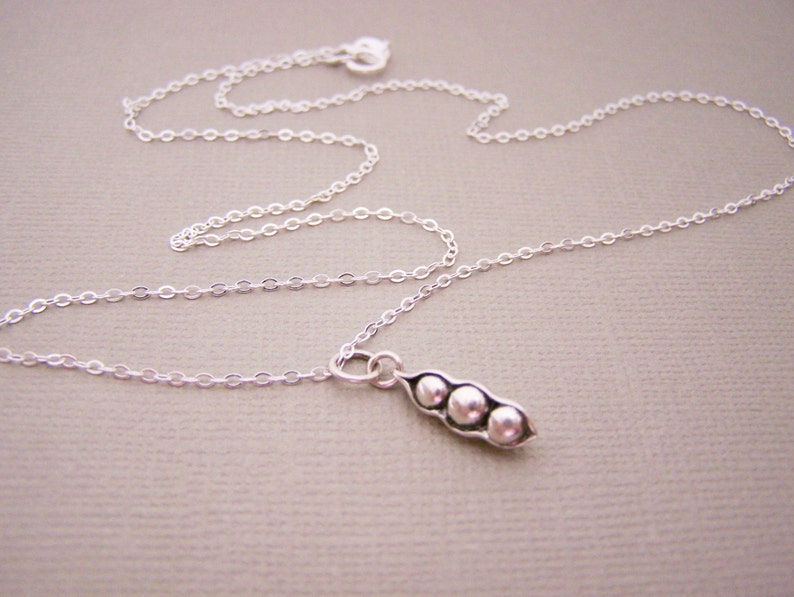 May include: A delicate silver necklace featuring a pea pod pendant with three small, round peas. The chain is thin and the pendant is detailed. The necklace is displayed on a neutral background.