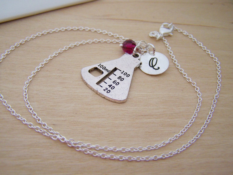 May include: A silver necklace with a beaker charm, a small pink bead, and a silver charm with the letter Q. The beaker charm has markings for 100 ml, 80 ml, 60 ml, 40 ml, and 20 ml.
