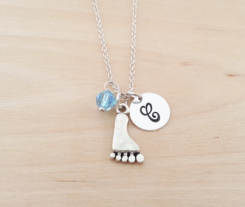 May include: Silver necklace with a charm of a footprint, a blue crystal bead, and a silver disc with the letter 'L' on it.