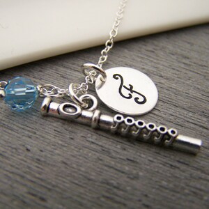 May include: A silver-colored necklace featuring a flute charm, a round pendant with a musical note, and a blue crystal bead. The necklace is displayed on a gray wooden surface.