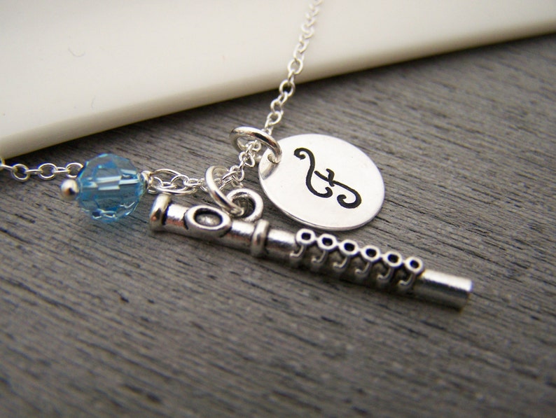 May include: A silver-colored necklace featuring a flute charm, a round pendant with a musical note, and a blue crystal bead. The necklace is displayed on a gray wooden surface.