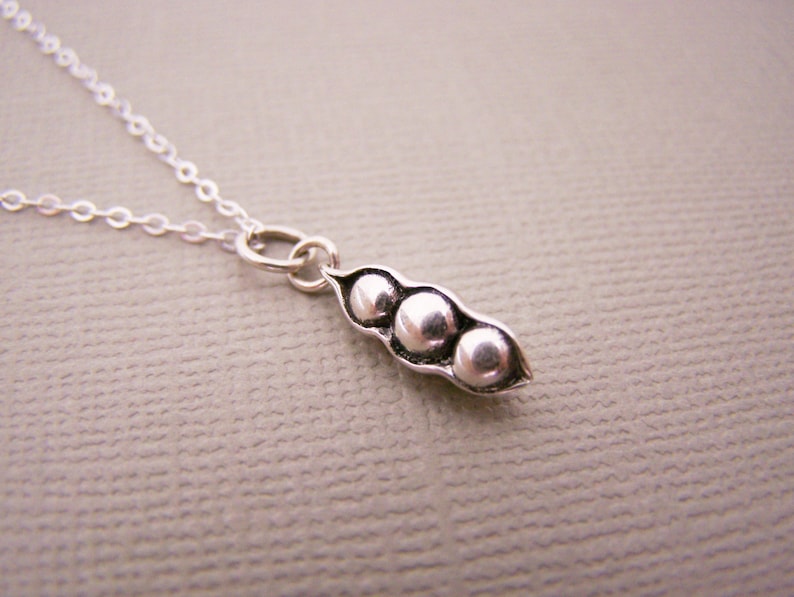May include: A silver necklace featuring a pendant shaped like a pea pod with three peas. The pendant is attached to a delicate silver chain. The pea pod pendant is approximately 0.75 inches long.