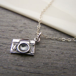 May include: A silver-colored camera pendant necklace. The pendant is a detailed miniature replica of a vintage camera. The necklace chain is delicate and silver. The camera pendant is approximately 0.75 inches wide.