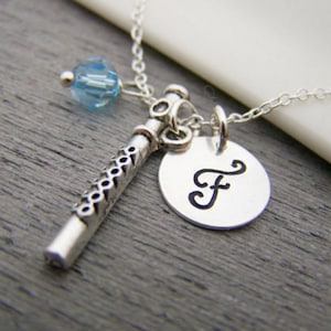 May include: Silver-toned necklace featuring a flute charm, a round pendant with the letter "F", and a light blue crystal bead. The flute charm has a detailed design. The necklace is on a gray surface.