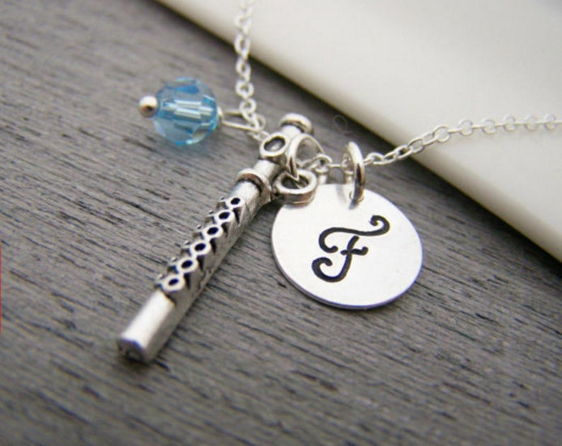 May include: Silver-toned necklace featuring a flute charm, a round pendant with the letter "F", and a light blue crystal bead. The flute charm has a detailed design. The necklace is on a gray surface.
