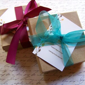 Puede incluir: Dos cajas de regalo envueltas en papel marr&oacute;n con cinta decorativa. Una caja est&aacute; coronada con una cinta azul turquesa y la otra con una cinta burdeos. La caja con la cinta azul turquesa tiene una tarjeta de visita adjunta con el texto "Design Studio, cyagmil@gmail.com, cydesignstudio.etsy.com".