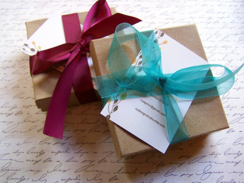 Puede incluir: Dos cajas de regalo envueltas en papel marr&oacute;n con cinta decorativa. Una caja est&aacute; coronada con una cinta azul turquesa y la otra con una cinta burdeos. La caja con la cinta azul turquesa tiene una tarjeta de visita adjunta con el texto "Design Studio, cyagmil@gmail.com, cydesignstudio.etsy.com".