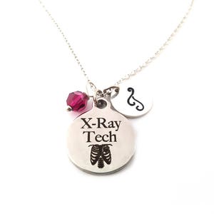May include: A silver necklace with a pendant that says "X-Ray Tech" with a rib cage graphic. A small, round charm with a medical symbol and a dark pink crystal bead are also attached to the chain.