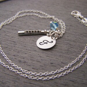 May include: Silver-toned necklace featuring a flute charm, a blue crystal bead, and a small, engraved oval pendant with a cursive letter. The chain is delicate and the clasp is visible.