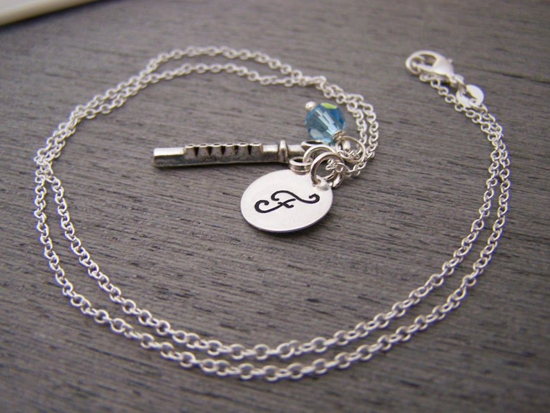 May include: Silver-toned necklace featuring a flute charm, a blue crystal bead, and a small, engraved oval pendant with a cursive letter. The chain is delicate and the clasp is visible.