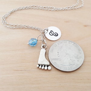 May include: Silver necklace featuring a footprint charm, a small blue crystal bead, and a silver disc with a cursive "&" symbol. The necklace is displayed on a wooden surface.
