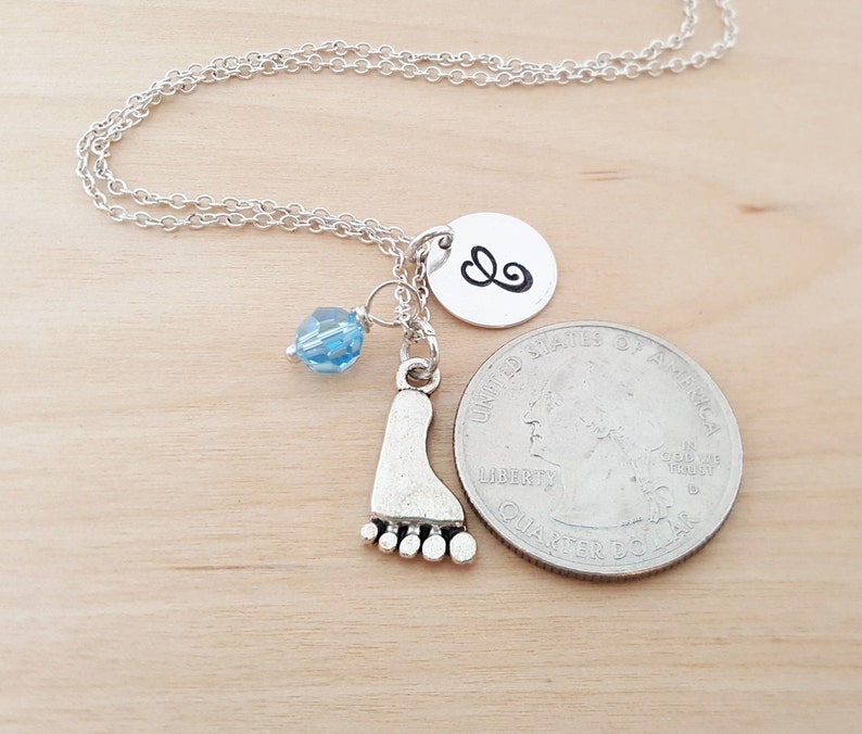 May include: Silver necklace featuring a footprint charm, a small blue crystal bead, and a silver disc with a cursive "&" symbol. The necklace is displayed on a wooden surface.