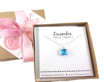 May include: A small gift box with a pink ribbon and a card inside. The card says "December - Blue Topaz" and has a silver necklace with a blue topaz pendant.