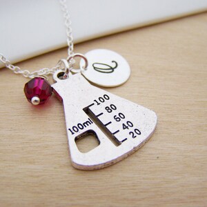 May include: Silver necklace with a beaker charm, a red crystal bead, and a silver initial charm. The beaker charm has a measurement scale from 20 to 100 ml.