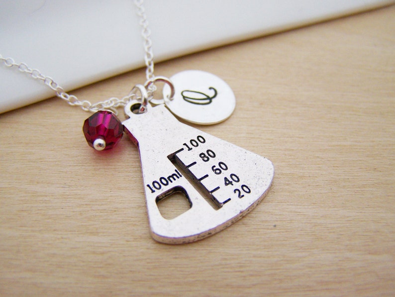 May include: Silver necklace with a beaker charm, a red crystal bead, and a silver initial charm. The beaker charm has a measurement scale from 20 to 100 ml.