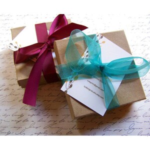 May include: Two small gift boxes wrapped in brown paper with a burgundy ribbon on one and a turquoise ribbon on the other. The boxes are sitting on a white surface with handwritten text.
