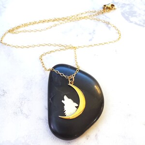May include: Gold necklace with a crescent moon pendant featuring a wolf silhouette. The pendant is on a black stone.