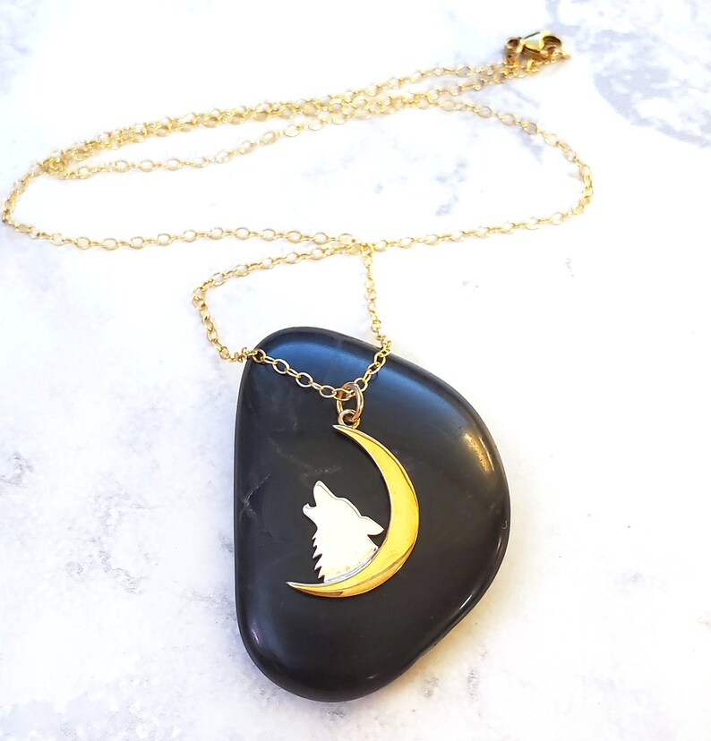 May include: Gold necklace with a crescent moon pendant featuring a wolf silhouette. The pendant is on a black stone.