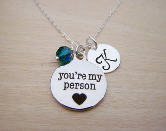 May include: Silver necklace with a round pendant that says "you're my person" with a small heart below the text. The necklace also has a small initial charm and a green crystal bead.