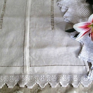 Op de afbeelding: Witte linnen theedoek met delicate gehaakte kant en geborduurde details. De handdoek is dubbelgevouwen en heeft een gegolfde rand.