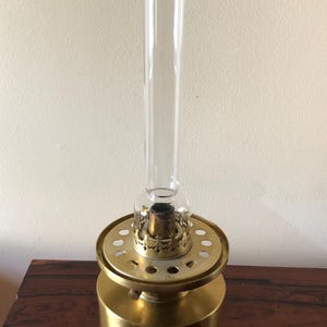 May include: A vintage brass oil lamp with a clear glass chimney. The lamp has a round base and a decorative top with a series of holes. The lamp is sitting on a dark wood surface.
