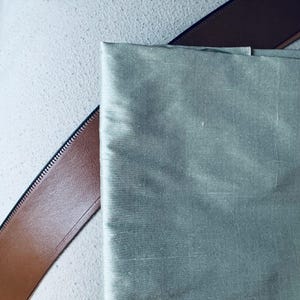 May include: A close-up of a piece of light green fabric with a smooth, silky texture. The fabric is lying on top of a brown leather strap.