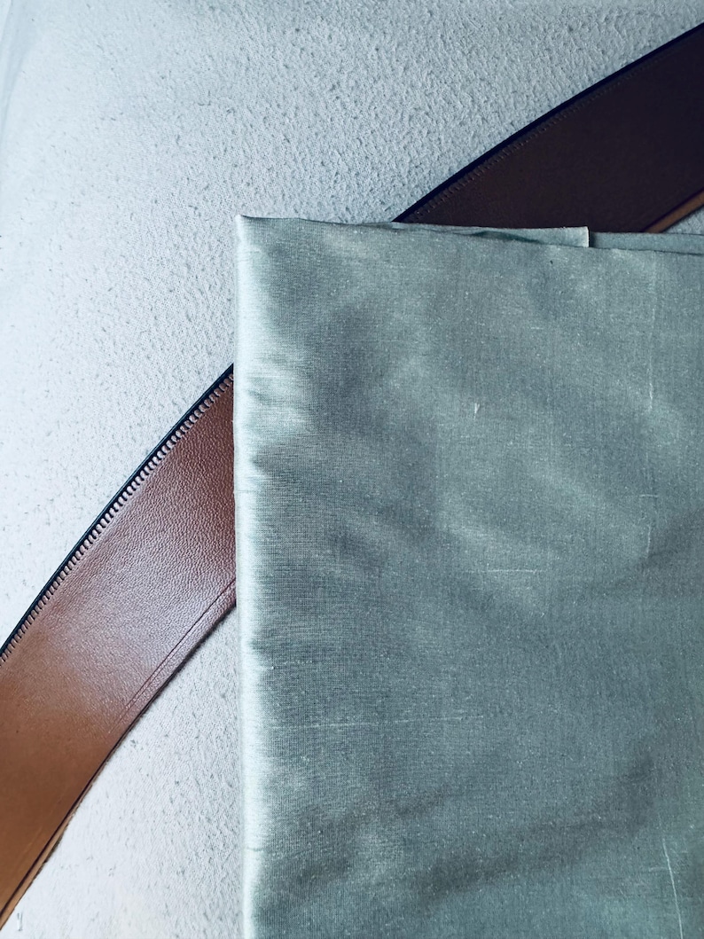 May include: A close-up of a piece of light green fabric with a smooth, silky texture. The fabric is lying on top of a brown leather strap.