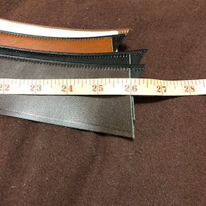 May include: Three strips of leather in different colors, brown, black, and white, with stitching around the edges. The leather strips are laid out on a brown fabric surface with a measuring tape showing 22 to 28 inches.