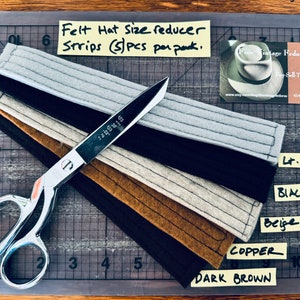 May include: Five felt hat size reducer strips in different colors: light grey, black, beige, copper, and dark brown. The strips are laid out on a cutting mat with a pair of scissors. The text "Felt Hat Size reducer Strips (5) pcs per pack" is written on a yellow sticky note.