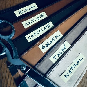 May include: Six leather straps in different colors, labeled with sticky notes: Black, Antique, Chocolate, Ginger, Taupe, and Natural. A pair of silver scissors is lying on top of the straps.