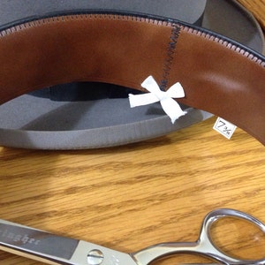 May include: A brown leather hat band with a white ribbon and a size tag that reads "7 1/8". A pair of silver scissors are in the foreground.
