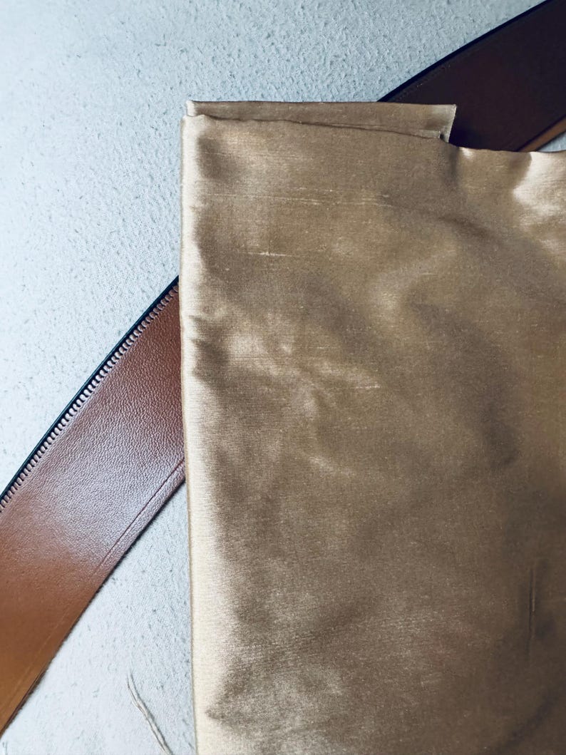 May include: A piece of gold fabric with a smooth, silky texture. The fabric is lying on top of a brown leather strap.