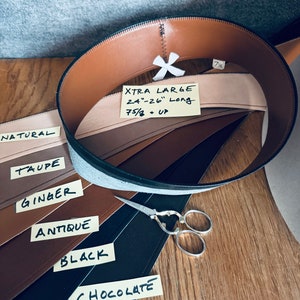May include: Leather straps in various colors, including natural, taupe, ginger, antique, black, and chocolate. The straps are labeled with their color names. A pair of scissors is also visible. The text "XTRA LARGE 24"-26" Long 7 5/8 + UP" is written on a yellow sticky note.