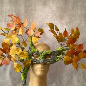 May include: A masquerade mask made of bark and moss with branches and leaves in shades of orange, yellow, and green.