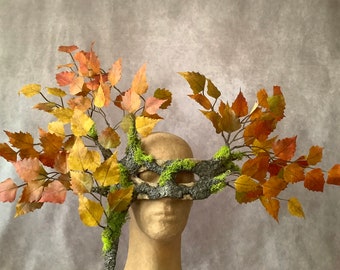 May include: A masquerade mask made of bark and moss with branches and leaves in shades of orange, yellow, and green.