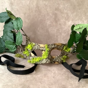May include: A green and brown masquerade mask with a wood and moss design. The mask has two eye holes and is attached to a black ribbon.