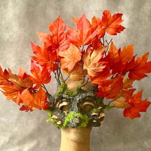 May include: A handcrafted mask featuring a face-like design with a mossy texture, adorned with small mushrooms and vibrant orange maple leaves. The leaves are arranged to create a crown-like effect, evoking an autumnal theme.