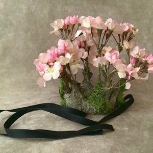May include: A crown made of moss and branches with pink flowers, tied with a black ribbon.