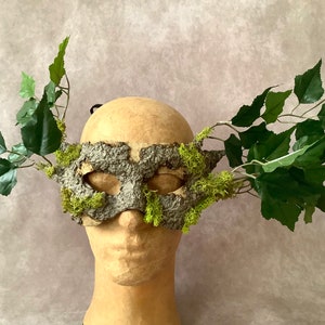 May include: A green and gray masquerade mask with moss and leaves attached to the sides. The mask is on a mannequin head.