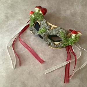 May include: A masquerade mask with a woodland theme. The mask is made of a light brown material and is decorated with moss, small red mushrooms, and white and red ribbons.