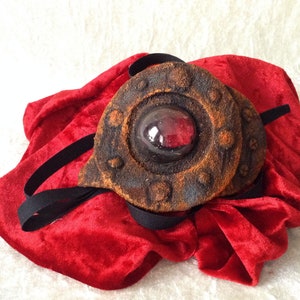 Peut inclure: Un masque pour les yeux de style steampunk avec un design circulaire brun vieilli et un grand centre rouge en forme de gemme. Le masque repose sur un tissu de velours rouge vif, avec un élastique noir.