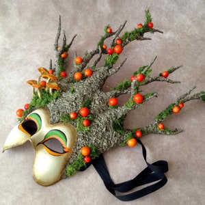 May include: A whimsical masquerade mask with a tree bark design, green moss, orange berries, and a black ribbon. The mask has a painted face with green and orange accents.