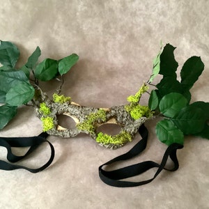 May include: A handmade masquerade mask made from tree bark and moss with green leaves attached. The mask has a black elastic strap.