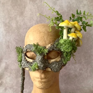 May include: A masquerade mask made of bark and moss with yellow mushrooms and green foliage attached to the top.