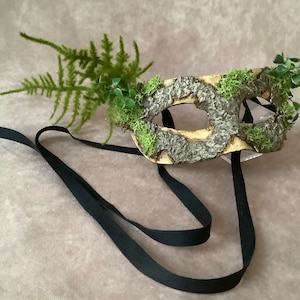 May include: A woodland-themed masquerade mask with a bark-like texture and green moss accents. The mask is attached to a black ribbon.