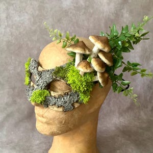 May include: A woodland-themed mask with a bark-like texture, adorned with green moss, faux mushrooms, and greenery. The mask is designed to be worn and is suitable for costume or decorative purposes.