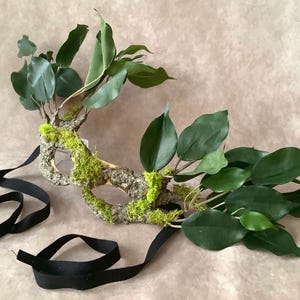 May include: A green and brown masquerade mask made of natural materials, with a leafy design and black ribbon ties.