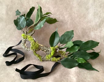 May include: A masquerade mask constructed from textured tree bark and vibrant green moss, adorned with clusters of dark green leaves extending from the sides. The mask features two eye openings and is secured with long, flat black fabric ribbons.