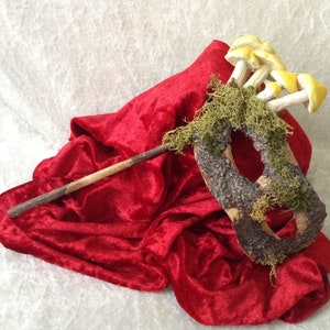 May include: A wooden mask with moss and yellow mushrooms attached to it. The mask is lying on a red velvet fabric.