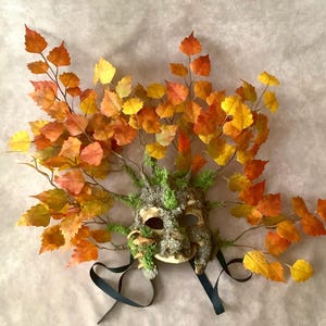May include: A handcrafted mask with a natural, earthy aesthetic. The mask features a textured, bark-like surface adorned with vibrant, autumn-colored leaves in shades of yellow, orange, and red. Dark ribbons hang from the mask's base.